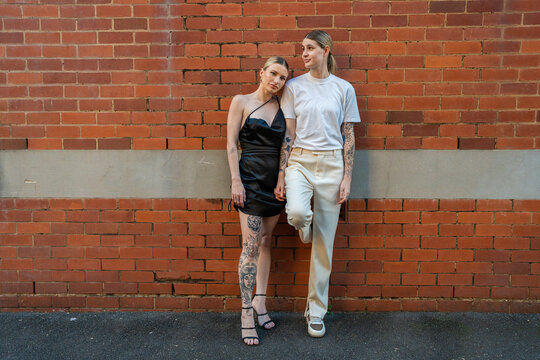 cool lesbian couple leaning against orange brick wall in street with serious expression