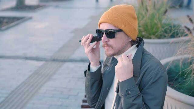Young blind man with smartphone sitting on bench in park in city, calling