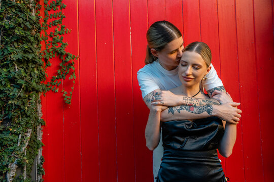 lesbian couple hugging and kiss on forehead with eyes closed in front of red wall 