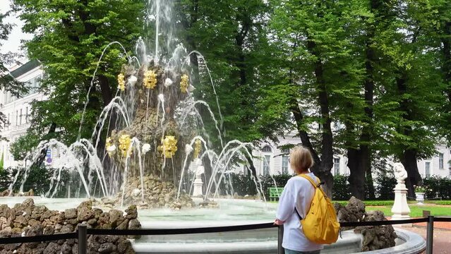 Young woman with a yellow backpack stands at a beautiful fountain complex, admires and calms down with the murmur and streams of water, a view from the back.