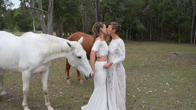 country lesbian brides kissing with horses at farm on wedding day
