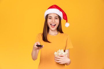 Young woman wearing Christmas hat with holding a red bucket of popcorns while the TV remote to...