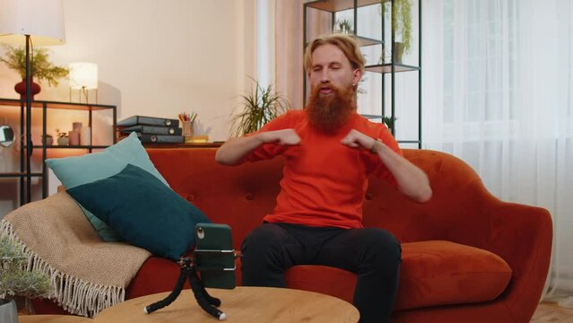 Portrait of caucasian bearded man blogger taking selfie on smartphone tripod, dancing, recording video vlog, communicating call online with subscribers. Young guy at home sitting on orange couch