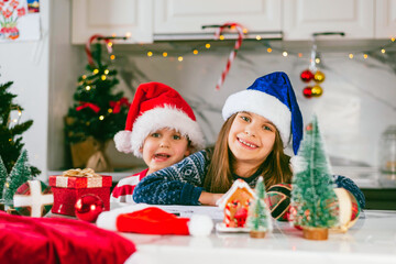 Fototapeta na wymiar Funny toddler boy and cute girl in a Santa hat is sitting in the kitchen with a decor for Christmas and looking at camera. Holidays, Traditions, baby food and health concept.