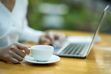 Young woman sitting in a coffee shop at a wooden table Drink coffee and use a laptop young women surfing the internet chat blogging woman holding a cup of coffee in a cafe.