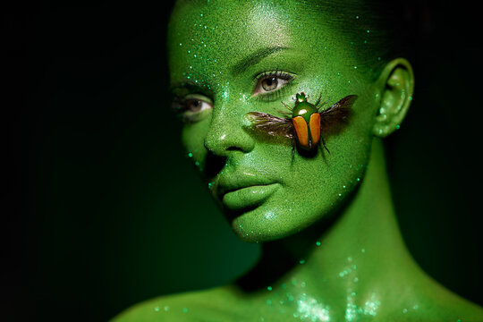 Fashion model woman skin face in bright sparkles with green beetle colorful neon lights, beautiful girl sexy lips. Glowing art green skin make-up. Glitter metallic shine silver green makeup