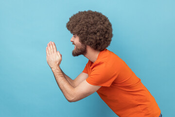 Side view of man with Afro hairstyle in orange T-shirt folding hands in prayer, closing eyes and...