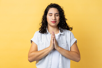 Portrait of peaceful calm relaxed woman with dark wavy hair standing with prayer gesture, keeping...