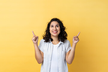 Portrait of satisfied delighted woman with dark wavy hair pointing up at empty place for ad content and expressing positive emotions. Indoor studio shot isolated on yellow background.