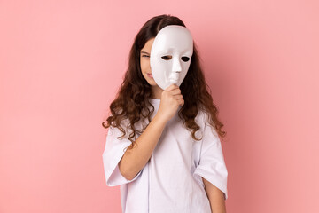 Portrait of dark haired little girl wearing white T-shirt covering face, standing with frowning...