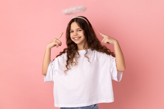 Portrait of cute little girl wearing white T-shirt pointing at holy nimbus over head, showing aureole and looking up with toothy smile. Indoor studio shot isolated on pink background.