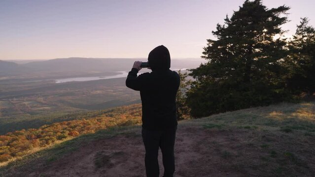 Unrecognizable Man with Black Jacket Standing on the Mountain Peak Facing to the Valley of Autumn Fall Foliage Trees Arkansas Taking Photo with His Smartphone