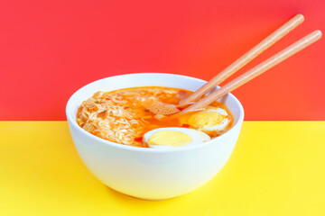 Bowl with instant noodle soup and eggs on red and yellow background close-up, side view.