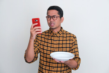 Adult Asian man looking to his handphone with unhappy expression while holding dinner plate