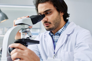 Modern male virologist in labcoat concentrating on study of new bacteria or virus while looking in microscope in clinical laboratory