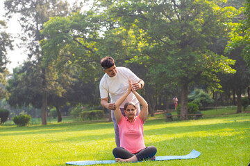 Indian male teacher or trainer teach yoga to female student in the park, Two people do stretching fitness exercise together outdoor, Sports and workout concept.