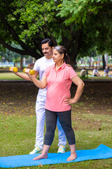 Indian trainer help a woman with dumbbell workout at park, working on arms strength, Asian athletic lady exercising outdoor with coach in nature, fitness concept.