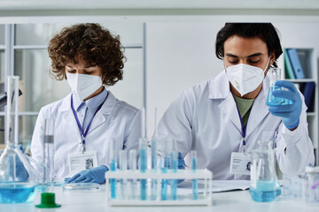 Fototapeta premium Two young clinicians in labcoats, respirators and gloves working with liquid chemical substances created in laboratory during trial