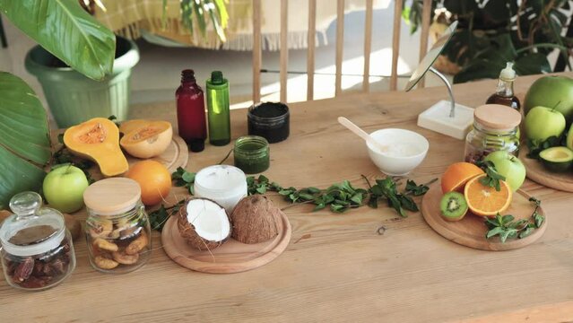 Various ingredients lying at wooden table for preparing natural cosmetics at home. Kiwi, orange, avocado, mint, coconut skin and hair care home spa. Bottle of oil, jar of mask, cup with cream.