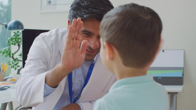Smiling doctor or GP in white coat meeting with mother and son in office for appointment giving boy high five - shot in slow motion