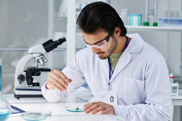 Fototapeta premium Young clinician in protective eyeglasses bending over workplace and dropping liquid substance into petri dish with bacteria