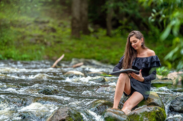 woman freelancer sitting on a rock in the river and using a laptop, travel or vacation work concept