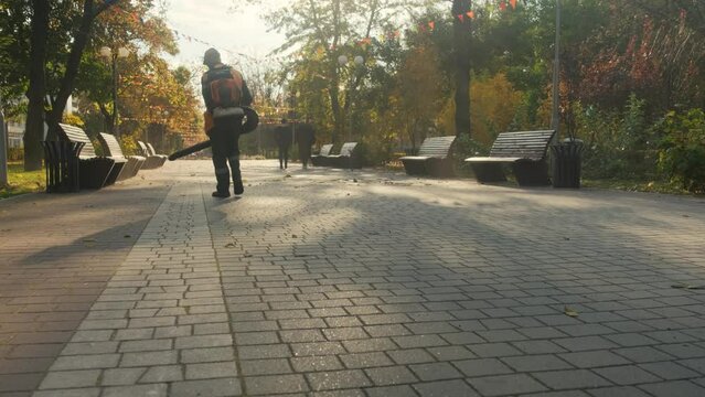 A man in a park with a blower removes dust and autumn leaves.