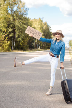 Vertical begging funny smiling blond woman autostop hitchhiking by raised leg sign, with plate anywhere carrying luggage