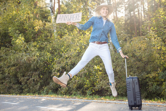 Active, funny, positive blond woman hitchhiker glad of car traffic, jumping in air, hold blank carton board with luggage