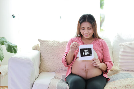 Happy smiling young asian pregnant woman resting and sitting on sofa in living room while showing ultrasound image. Expectant mother preparing and waiting for baby birth during pregnancy.