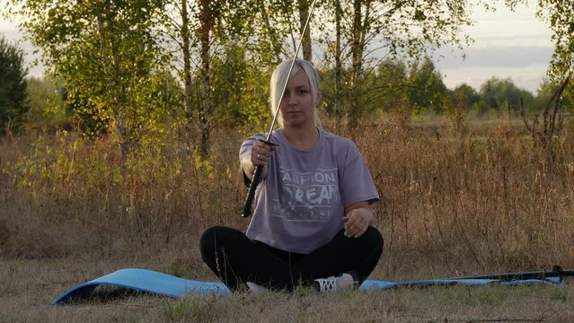 A Woman Sits On A Rug And Gives A Katana From A Scabbard. A Blond Woman Pulls Out A Katana And Holds It In Front Of Her, Then Aiming It At The Camera. Concept Woman With Katana.