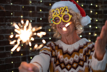Handsome blonde senior woman with funny glasses and winter sweater holding sparkler light, elderly...