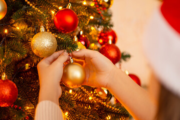 Girl decorating Christmas tree. Focus on hands of a girl hanging golden and red baubles to the branches of Christmas tree