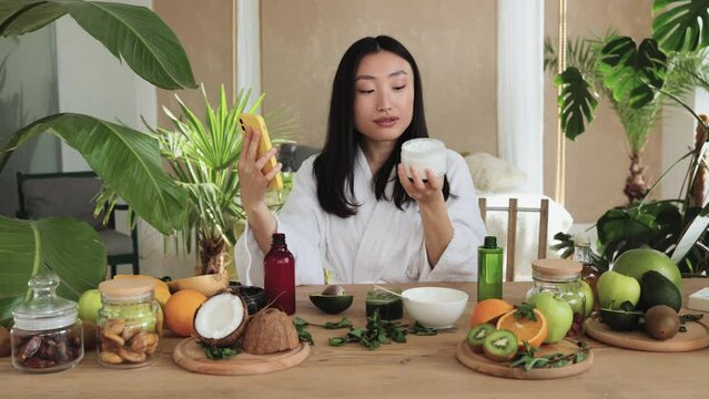 Asian brunette shoulder length hair female social media influencer in bathrobe streaming and introducing beauty products to audience with phone holding glass jar with cream, skin care concet.