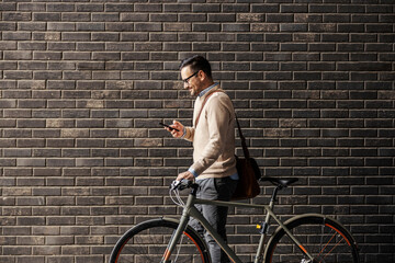 A smart casual businessman is pushing his bicycle and passing by the wall while texting a message...