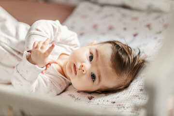 A cute baby girl with blue eyes is looking at the camera while lying in a crib. - obrazy, fototapety, plakaty