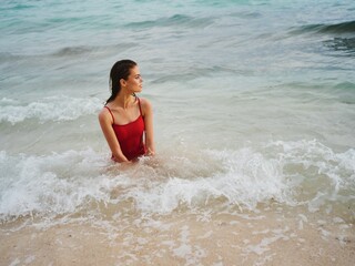 Fototapeta premium Sexy woman in a red swimsuit with an athletic body stands in the water in the ocean and poses looking out at the horizon