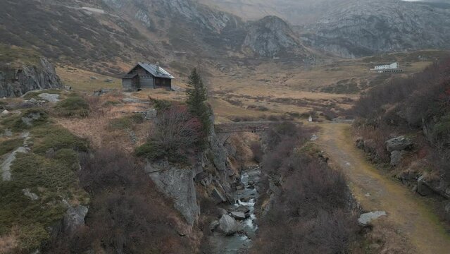 Old wooden hut on the pass road to the Nufenen Pass in Switzerland. A stony river bed flows down the valley and a hiking trail leads down the side - Aerial Shot