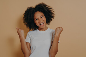 Young euphoric woman clenching fists and celebrating achievement, isolated on yellow background