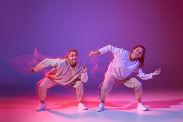 Fototapeta na wymiar Portrait of two young girls dancing hip-hop isolated over gradient purple background in neon with mixed light