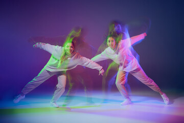 Fototapeta na wymiar Portrait of young girls dancing hip-hop isolated over gradient blue purple background in neon with mixed light. Friendship