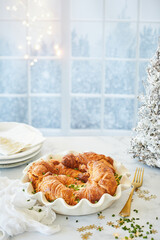Christmas breakfast with baked croissants filled with ham and cheese, served in a ceramic baking dish. Festive holiday setting with snow-covered window, golden fork, and winter decorations.