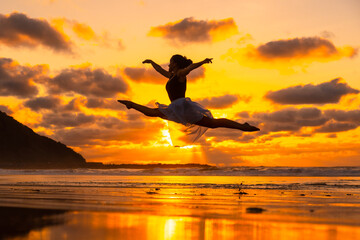 Young dancer on the beach at sunset performing a jump with the sea in the background, with the sun...
