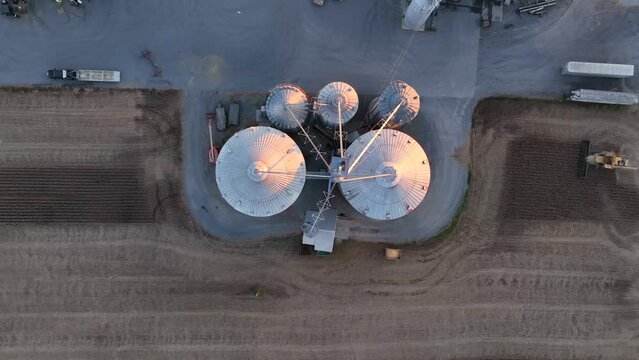 Top down aerial of combine harvesting beside grain elevator bins at golden hour.