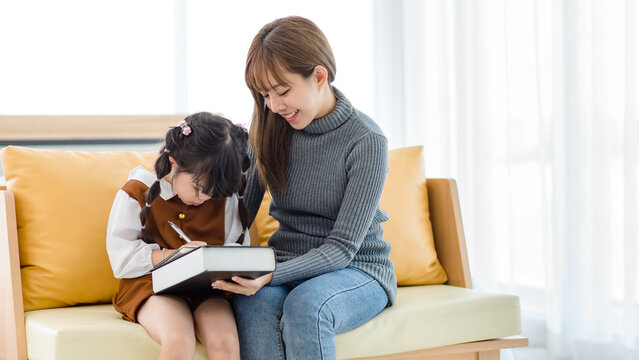 Millennial Asian young female teenager mother nanny babysitter in casual outfit sitting on sofa smiling helping teaching homework to little cute preschooler daughter girl writing drawing with pen