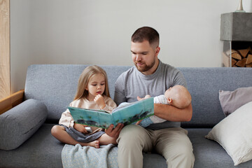 Young single dad with two children sitting on couch at home, reading book to his daughter and...