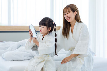 Closeup shot of Millennial Asian happy cheerful little cute preschooler daughter girl wearing white bathrobe after bath looking to mirror in hands setting hairstyle with unrecognizable unknown mother