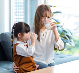 Millennial Asian young pretty female teenager mother nanny babysitter in casual outfit sitting on sofa smiling holding serving delicious milk glass to little cute preschooler daughter girl drinking