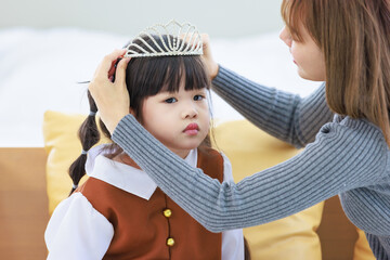 Closeup shot of Millennial Asian pretty young female teenager mother nanny babysitter role playing smiling wearing diamond princess crown on happy cheerful little cute preschooler daughter girl head © Bangkok Click Studio