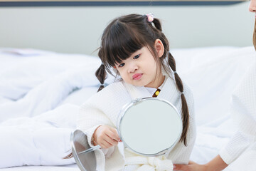 Closeup shot of Millennial Asian happy cheerful little cute preschooler daughter girl wearing white bathrobe after bath looking to mirror in hands setting hairstyle with unrecognizable unknown mother